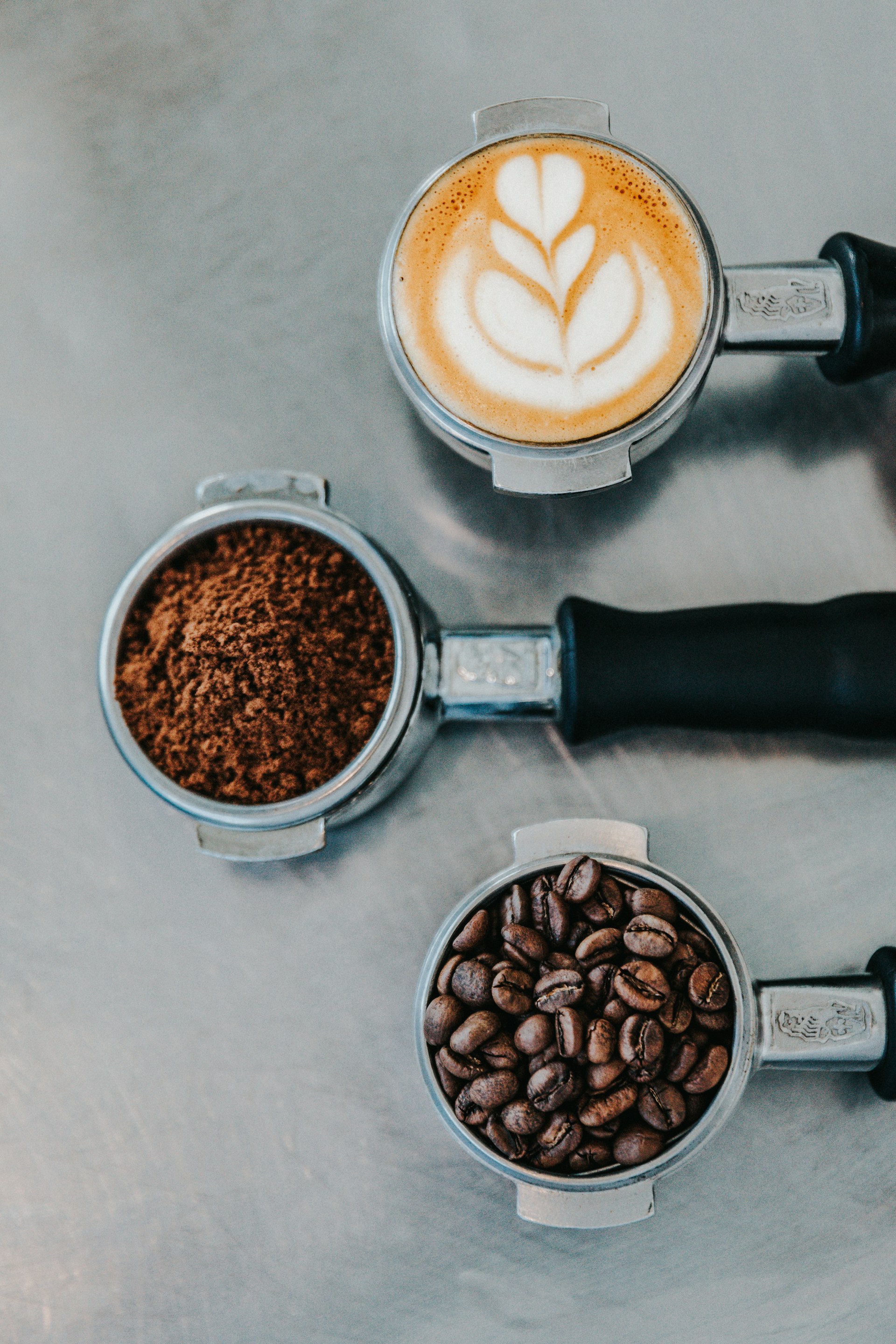 Three portafilters which are contain latte art, ground coffee, and whole coffee beans arranged vertically are there